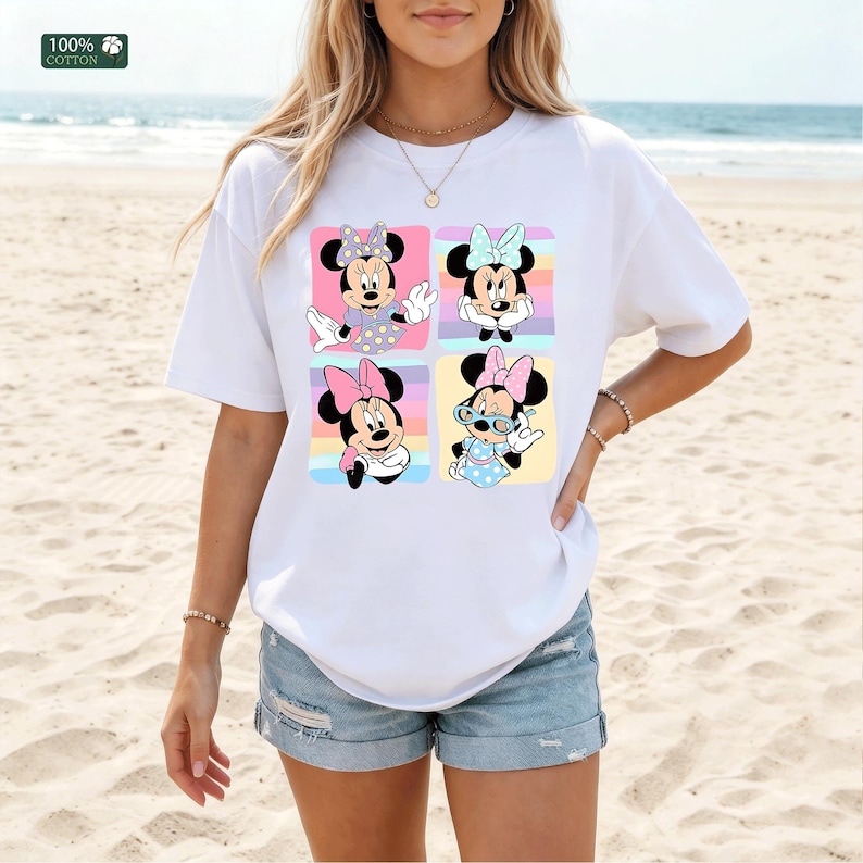May include: A white short-sleeved t-shirt made of 100% cotton featuring a graphic print of four distinct squares, each displaying a different illustration of Minnie Mouse in various poses and outfits against colorful pastel striped backgrounds.
