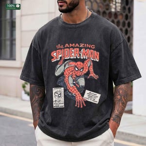 May include: A charcoal gray short-sleeve t-shirt featuring a distressed graphic of Spider-Man in a crouching pose. The text reads The Amazing Spider-Man, Peter Parker Alias Spider-Man, and Another Spectacular Smash-Hit From The House Of Ideas.