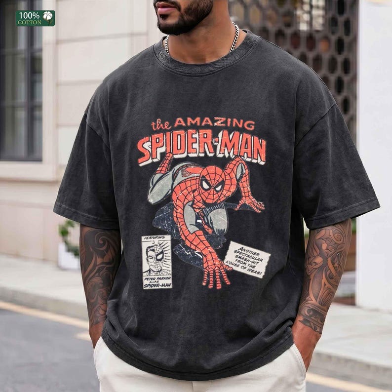 May include: A charcoal gray short-sleeve t-shirt featuring a distressed graphic of Spider-Man in a crouching pose. The text reads The Amazing Spider-Man, Peter Parker Alias Spider-Man, and Another Spectacular Smash-Hit From The House Of Ideas.