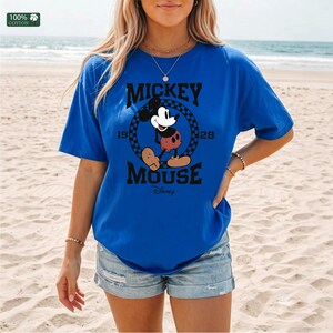 Peut inclure: Un t-shirt bleu vif &agrave; manches courtes avec un motif noir de Mickey Mouse dans un cercle &agrave; damier. Le texte indique MICKEY MOUSE 1928 et Disney. Une petite &eacute;tiquette carr&eacute;e en haut &agrave; gauche indique 100% COTTON. Le t-shirt a un col rond.