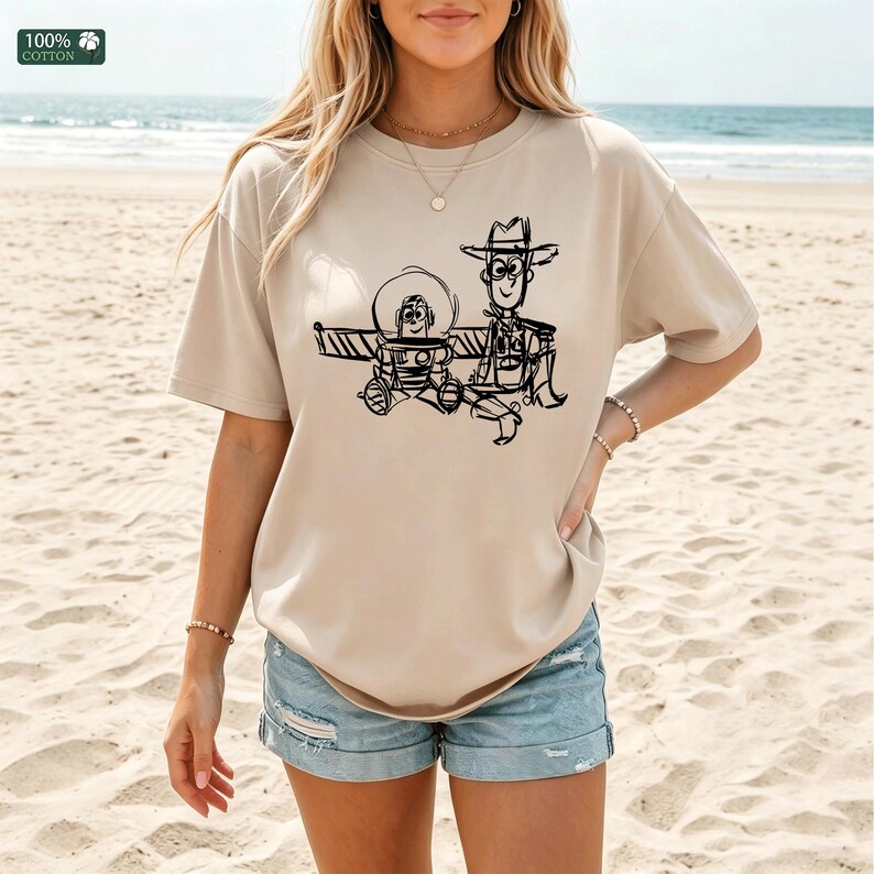 May include: A beige short-sleeved t-shirt featuring a black line-art graphic of two cartoon characters, one in a space helmet and one wearing a cowboy hat. The shirt has a crew neckline and a small green tag in the corner labeled 100% cotton.