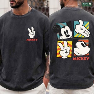May include: Dark gray t-shirt featuring Mickey Mouse. The front displays a white peace sign and the word "MICKEY" in red. The back showcases Mickey's face and hands in a four-square design with the word "MICKEY" in red. Made of 100% cotton.