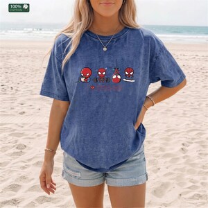 May include: A blue acid-wash t-shirt featuring a graphic print of four cartoon Spider-Man characters performing various activities including eating, lifting weights, hanging from a web, and skateboarding, with a small red heart icon.