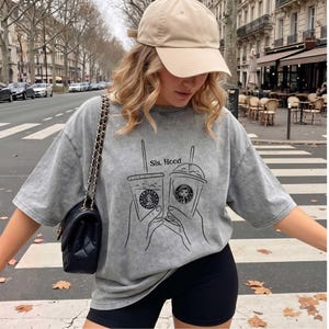 Puede incluir: Una camiseta gris con efecto lavado al &aacute;cido que presenta un gr&aacute;fico de l&iacute;neas negras de dos manos sosteniendo tazas de caf&eacute; con el texto Sis. Hood. Se combina con una gorra de b&eacute;isbol beige y un bolso de hombro negro acolchado con cadena dorada.