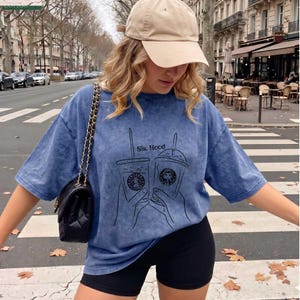 Puede incluir: Una camiseta azul con efecto lavado &aacute;cido que presenta un gr&aacute;fico de l&iacute;neas negras de dos manos sosteniendo vasos de caf&eacute; helado con el texto Sis. Hood. Se combina con pantalones cortos de ciclista negros, una gorra beige y un bolso acolchado.
