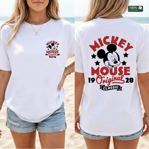 May include: White t-shirt featuring a Mickey Mouse graphic on the front and back. The front graphic includes Mickey's face, stars, and text. The back graphic is larger, with Mickey's face, stars, and the words "MICKEY MOUSE 19 Original 28 CLASSIC".