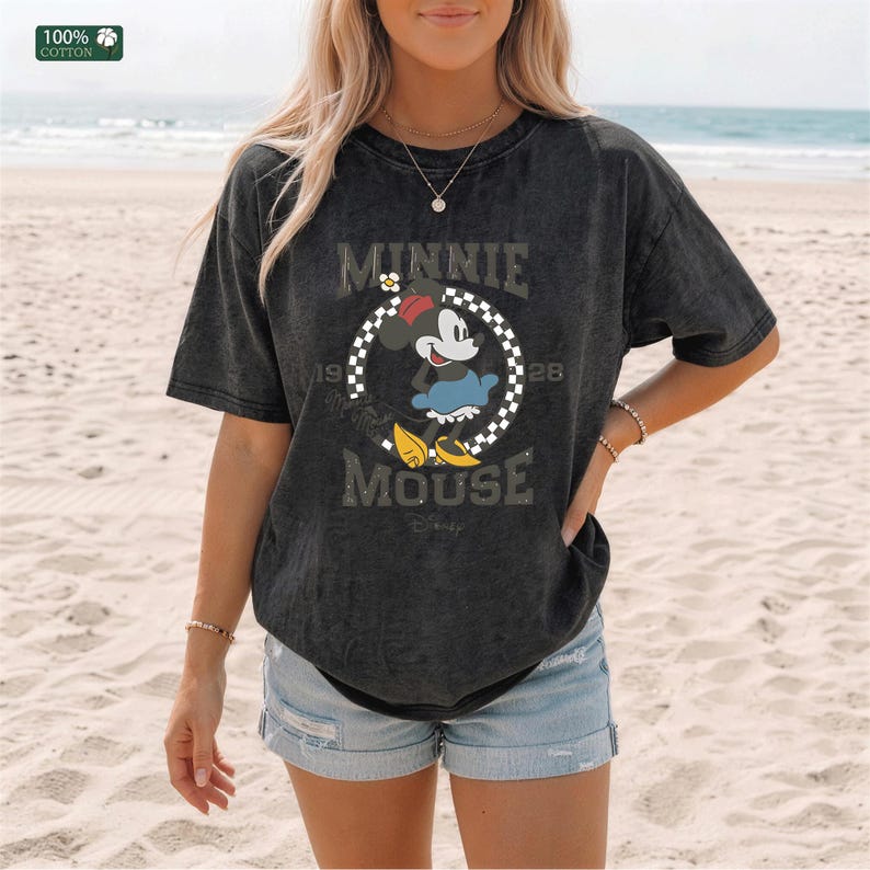 May include: A black, short-sleeved t-shirt featuring a distressed graphic of Minnie Mouse inside a checkered circle, with the text "MINNIE MOUSE" and "1928". The shirt is made of 100% cotton.