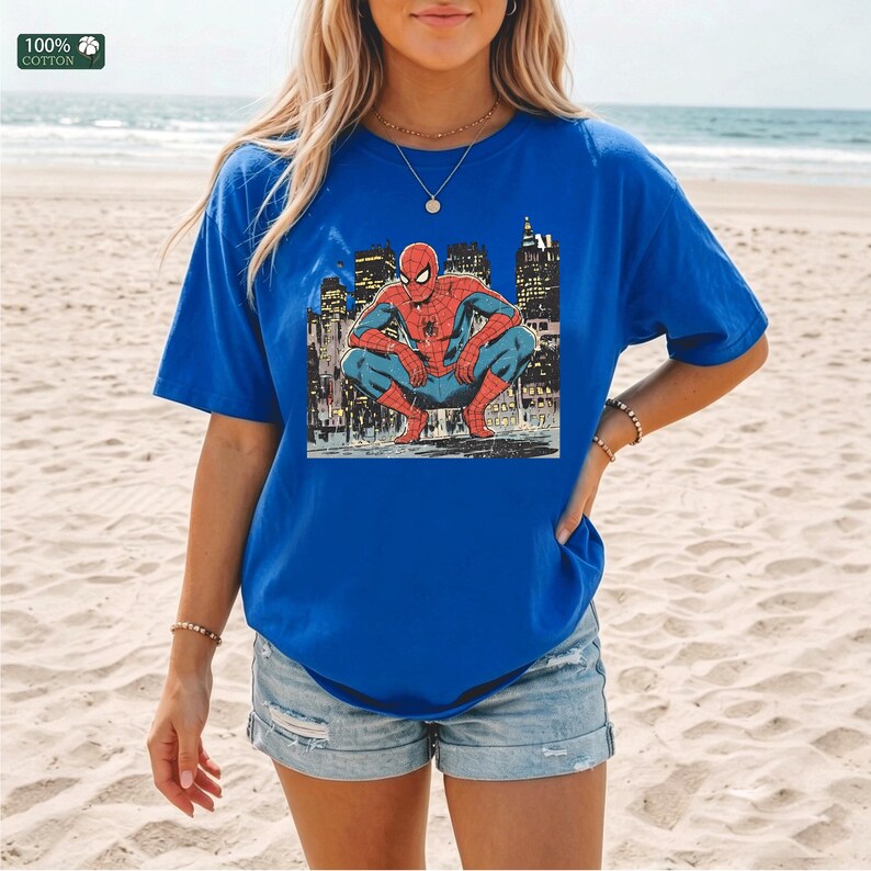 May include: A royal blue short-sleeve t-shirt featuring a graphic print of the superhero Spider-Man crouching in front of a city skyline. The shirt has a crew neckline and a small square label in the top left corner that reads 100% COTTON.