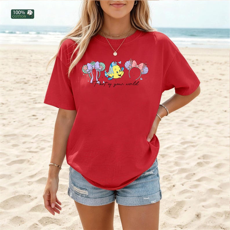 May include: A red short-sleeved t-shirt featuring a graphic of two pairs of mouse ears and a yellow fish character. The text "Part of your world" is printed in black cursive script below the graphic. A small label in the corner reads 100% cotton.