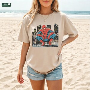 May include: A beige short-sleeved t-shirt featuring a graphic print of the superhero Spider-Man in a crouching position against a dark city skyline background. The shirt has a crew neckline and a relaxed fit, with a small 100% cotton label visible.