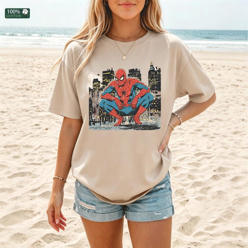 May include: A beige short-sleeved t-shirt featuring a graphic print of the superhero Spider-Man in a crouching position against a dark city skyline background. The shirt has a crew neckline and a relaxed fit, with a small 100% cotton label visible.