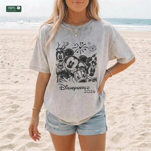 Puede incluir: Camiseta gris claro con un boceto negro de personajes de Disney, incluyendo Mickey Mouse, Minnie Mouse, Donald Duck y Goofy. El texto "Disney World 2026" est&aacute; debajo. La camiseta es 100% algod&oacute;n.
