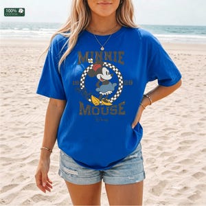 May include: Royal blue t-shirt featuring a Minnie Mouse graphic. The design includes the text "Minnie Mouse" and the years "1928" and "28". The shirt is made of 100% cotton.