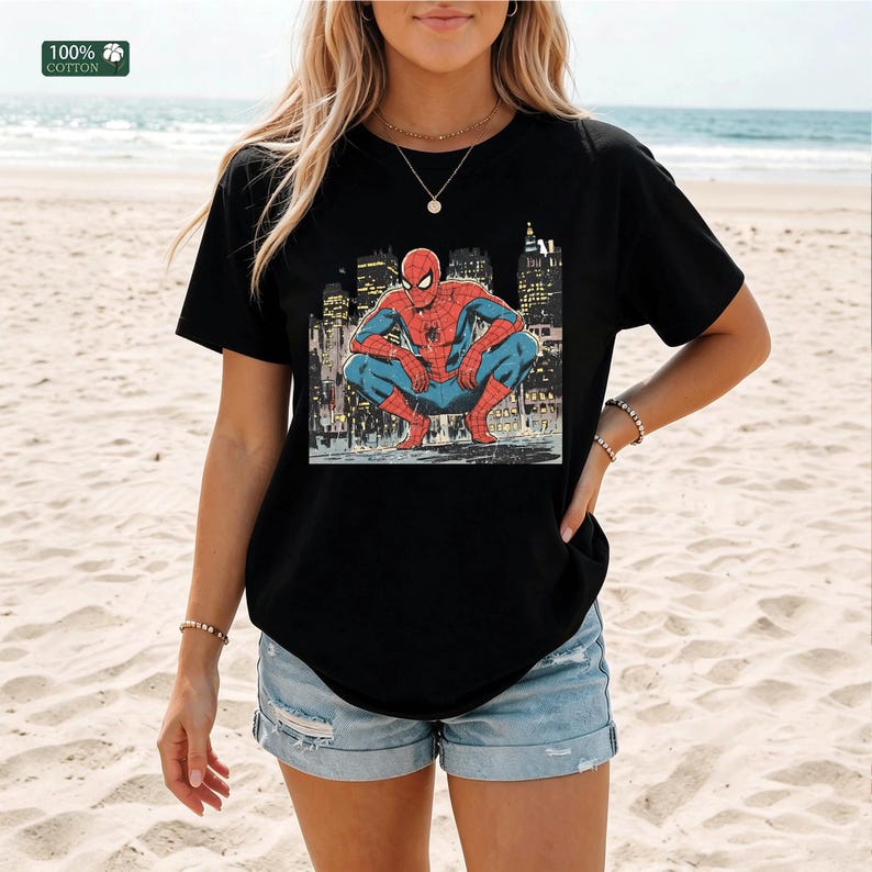 May include: A black short-sleeve t-shirt featuring a graphic print of the superhero Spider-Man in a crouching position against a city skyline background. The shirt includes a small green square label in the corner that reads 100% COTTON.