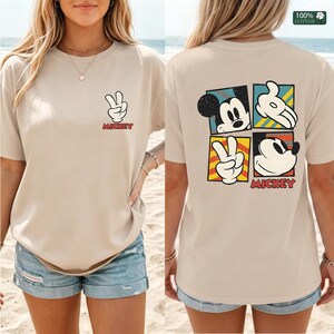 May include: Beige t-shirt featuring a Mickey Mouse peace sign graphic on the front and a four-panel design on the back. The back design includes Mickey's face, hands, and peace sign, with the word "Mickey" in red.