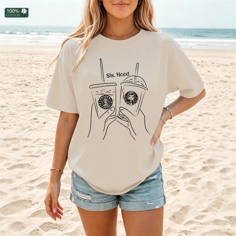 Puede incluir: Una camiseta beige de manga corta con un gr&aacute;fico de l&iacute;neas negras de dos manos sosteniendo vasos de caf&eacute; helado con pajitas. El texto "Sis. Hood" est&aacute; impreso sobre los vasos. Una peque&ntilde;a etiqueta verde "100% COTTON" es visible arriba a la izquierda.