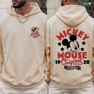 May include: Cream-colored hoodie featuring a front and back graphic of Mickey Mouse. The front graphic includes the text "Mickey Mouse 19 Original 28 Classic". The back graphic is a larger version of the same design. Made of 100% cotton.