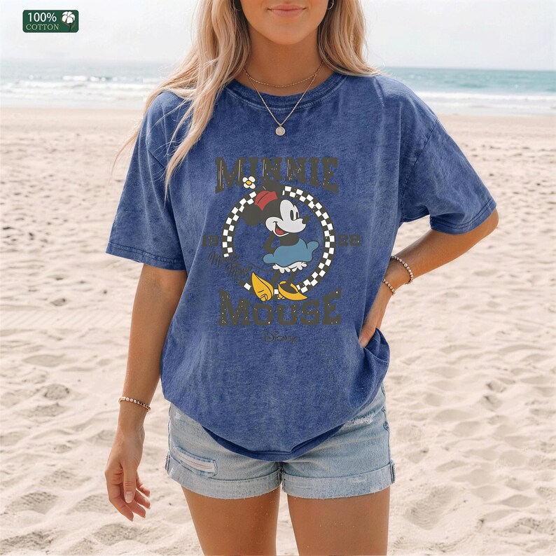 May include: Blue t-shirt with a graphic of Minnie Mouse in a circle with text. The shirt is made of 100% cotton and has a vintage wash. The text on the shirt says "Minnie Mouse". The shirt is worn with denim shorts.