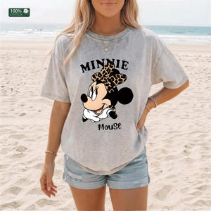 May include: A light gray t-shirt featuring a graphic of Minnie Mouse with a leopard print bow. The text "MINNIE" is above the character, and "Mouse" is below. The shirt is made of 100% cotton.