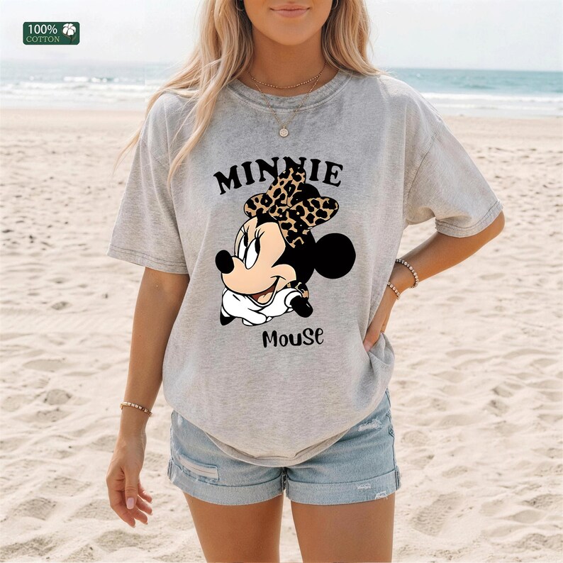 May include: A light gray t-shirt featuring a graphic of Minnie Mouse with a leopard print bow. The text "MINNIE" is above the character, and "Mouse" is below. The shirt is made of 100% cotton.