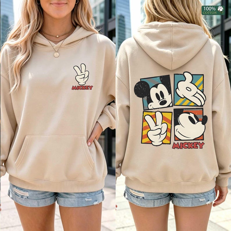 May include: Beige hoodie featuring Mickey Mouse graphics. The front displays a peace sign hand gesture with the word "Mickey" in red. The back showcases Mickey Mouse in four colorful squares, with "Mickey" at the bottom. Made of 100% cotton.