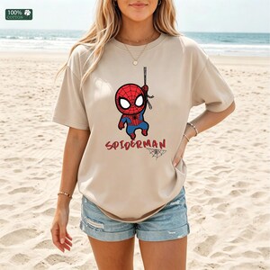 Op de afbeelding: Beige T-shirt met een cartoon Spider-Man afbeelding. Het ontwerp toont Spider-Man die aan een web hangt, met het woord "SPIDERMAN" in het rood eronder. Het shirt is gemaakt van 100% katoen.