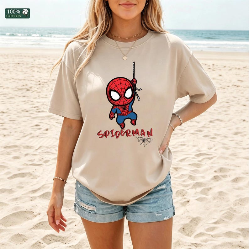 Op de afbeelding: Beige T-shirt met een cartoon Spider-Man afbeelding. Het ontwerp toont Spider-Man die aan een web hangt, met het woord "SPIDERMAN" in het rood eronder. Het shirt is gemaakt van 100% katoen.