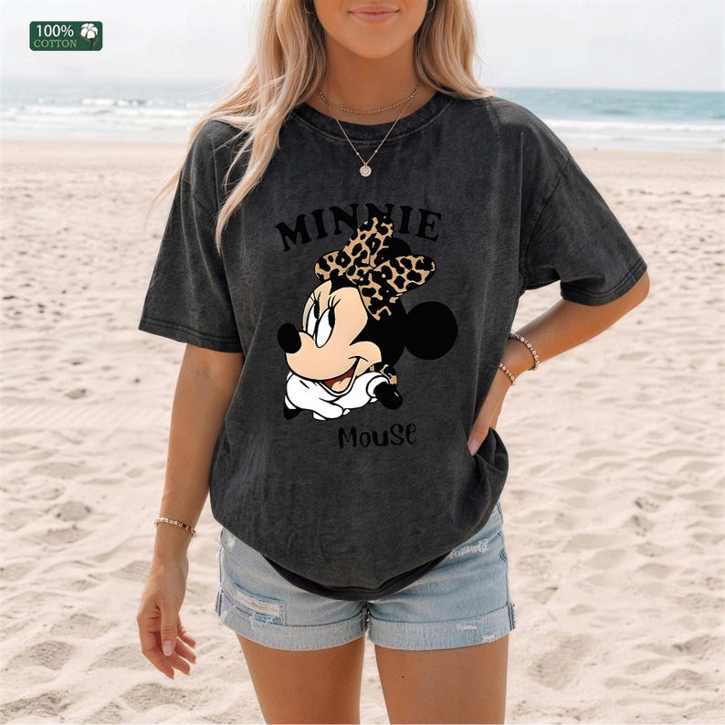 May include: Dark gray t-shirt featuring a Minnie Mouse graphic. The design includes the text "MINNIE MOUSE" above and below the character. The shirt is made of 100% cotton and has a relaxed fit.