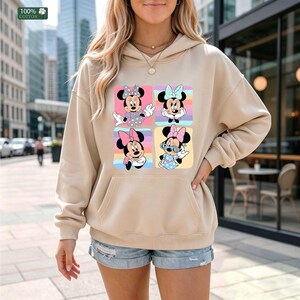 May include: A beige hooded sweatshirt featuring a graphic print of four squares, each containing a different illustration of Minnie Mouse in various outfits and poses. The hoodie has long sleeves, a front kangaroo pocket, and a 100% cotton label.