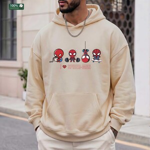 May include: A beige hooded sweatshirt featuring a graphic of four cartoon Spider-Man characters in various activities, including eating, lifting weights, hanging, and skateboarding, with the text I heart SPIDER-MAN printed below the figures.