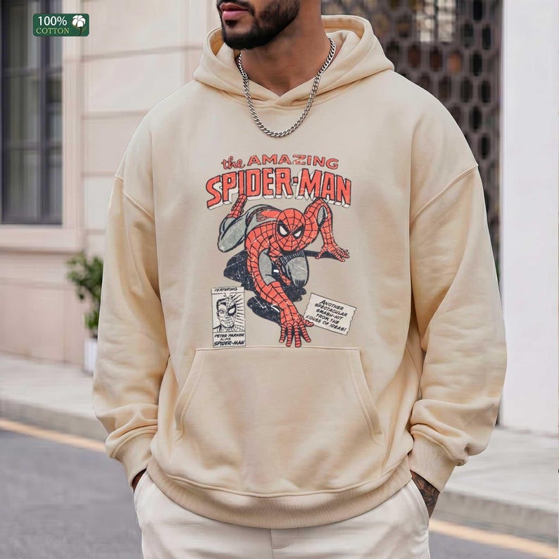 May include: A beige hooded sweatshirt featuring a graphic print of Spider-Man in a red and blue suit. The text reads The Amazing Spider-Man, Peter Parker, and Another Spectacular Smash-Hit from the Pages of Ideas. A 100% cotton label is visible.
