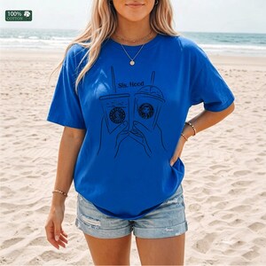Puede incluir: Una camiseta azul brillante de manga corta con un gr&aacute;fico de l&iacute;neas negras de dos manos sosteniendo vasos de caf&eacute; helado. El texto "Sis. Hood" est&aacute; impreso sobre los vasos. Una peque&ntilde;a etiqueta cuadrada indica "100% COTTON".