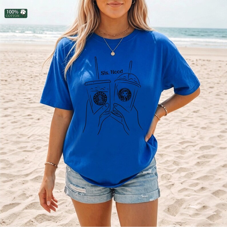 Puede incluir: Una camiseta azul brillante de manga corta con un gr&aacute;fico de l&iacute;neas negras de dos manos sosteniendo vasos de caf&eacute; helado. El texto "Sis. Hood" est&aacute; impreso sobre los vasos. Una peque&ntilde;a etiqueta cuadrada indica "100% COTTON".