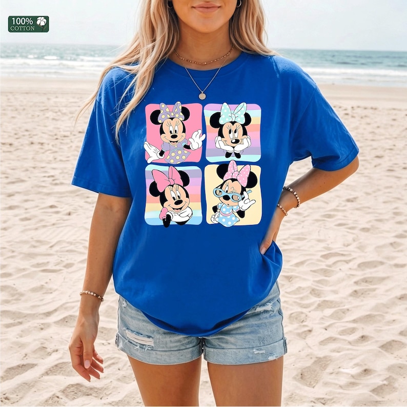 May include: A royal blue short-sleeve t-shirt featuring a graphic print of four squares, each containing a different illustration of Minnie Mouse in various poses and outfits. The top left corner includes a small square label stating 100% cotton.