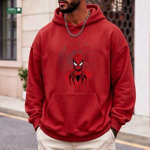 May include: Red hooded sweatshirt featuring a Spider-Man graphic. The graphic includes a detailed illustration of Spider-Man's face and costume, set against a web design. The sweatshirt has a front pocket and a drawstring hood. The fabric is 100% cotton.