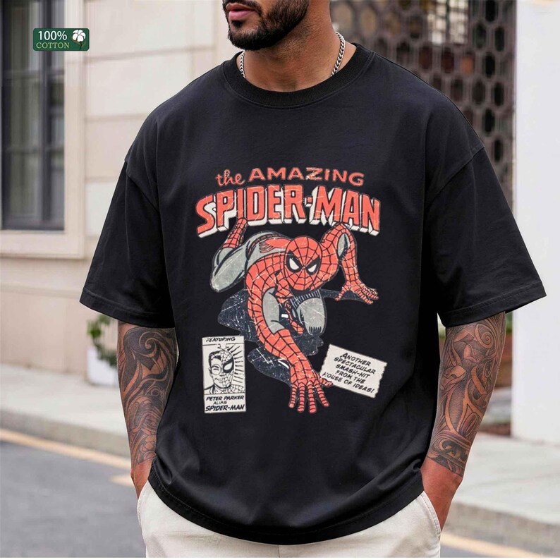 May include: A black short-sleeve t-shirt featuring a vintage-style graphic of Spider-Man in a crouching pose. The text reads "the AMAZING SPIDER-MAN" and includes a small box with "PETER PARKER ALIAS SPIDER-MAN" and a note about a smash-hit.