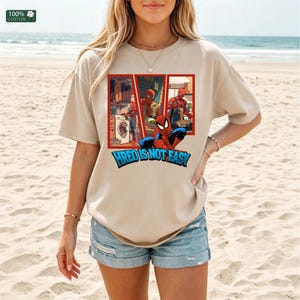 May include: A beige short-sleeved t-shirt featuring a graphic print of the superhero Spider-Man performing household chores like laundry and cooking. Below the graphic, the text reads HREO IS NOT EASY in bold blue letters with a black outline.