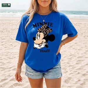 May include: Royal blue t-shirt featuring a Minnie Mouse graphic. The graphic includes the text "MINNIE Mouse" with Minnie wearing a leopard print bow. The shirt is made of 100% cotton.