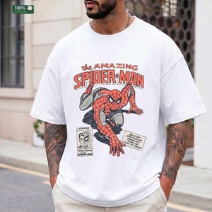 May include: A white short-sleeved t-shirt featuring a vintage-style graphic of Spider-Man in a crouching pose. The text reads The Amazing Spider-Man, Peter Parker Alias Spider-Man, and Another Spectacular Smash-Hit From The House Of Ideas.