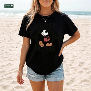 Peut inclure: Un t-shirt noir &agrave; manches courtes avec un motif de Mickey Mouse au centre. Le design inclut le texte Mickey Mouse et l'ann&eacute;e 1928. Une petite &eacute;tiquette carr&eacute;e verte dans le coin sup&eacute;rieur gauche affiche le texte 100% Cotton.