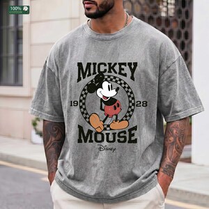 Peut inclure: Un t-shirt gris &agrave; manches courtes avec un motif de Mickey Mouse &agrave; l'int&eacute;rieur d'un cercle &agrave; damier noir et blanc. Le texte indique MICKEY MOUSE, 1928 et Disney. Le t-shirt pr&eacute;sente un aspect d&eacute;lav&eacute; et us&eacute; avec un col rond.