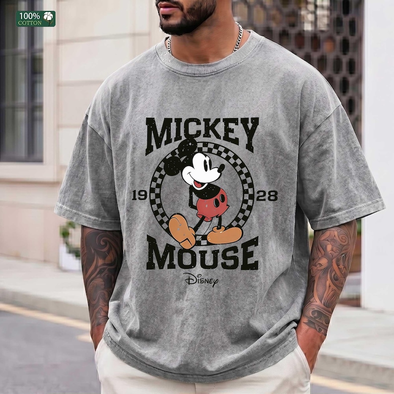 Peut inclure: Un t-shirt gris &agrave; manches courtes avec un motif de Mickey Mouse &agrave; l'int&eacute;rieur d'un cercle &agrave; damier noir et blanc. Le texte indique MICKEY MOUSE, 1928 et Disney. Le t-shirt pr&eacute;sente un aspect d&eacute;lav&eacute; et us&eacute; avec un col rond.