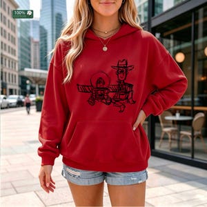 May include: A red long-sleeve hooded sweatshirt featuring a black line-art graphic of two cartoon characters, one in a space suit and one in a cowboy outfit. The hoodie includes a front kangaroo pocket and a 100% cotton label in the corner.