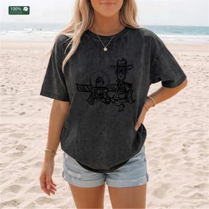 May include: A charcoal gray short-sleeve t-shirt made of 100% cotton, featuring a black line-art graphic of two cartoon characters, one wearing a space helmet and the other a cowboy hat, printed on the front center of the garment.
