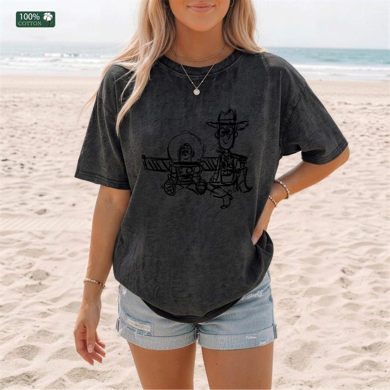 May include: A charcoal gray short-sleeve t-shirt made of 100% cotton, featuring a black line-art graphic of two cartoon characters, one wearing a space helmet and the other a cowboy hat, printed on the front center of the garment.