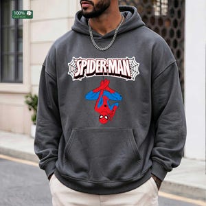 May include: A charcoal grey hoodie with a Spider-Man graphic. The graphic shows Spider-Man hanging upside down, with the word "SPIDER-MAN" in a white webbed design. The hoodie has a front pocket and a drawstring hood. The tag reads "100% cotton."