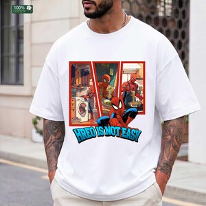 May include: A white short-sleeved t-shirt featuring a graphic print of the character Spider-Man in various domestic settings, including doing laundry and cooking. Below the graphic, the text reads HREO IS NOT EASY in bold blue letters.