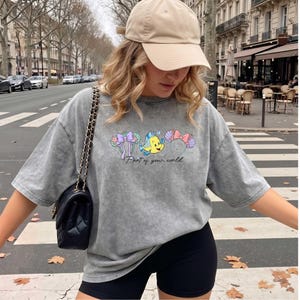 May include: A grey acid-wash t-shirt featuring a graphic of two pairs of mouse ears and a yellow fish character, with the text "Part of your world" printed below. The shirt has short sleeves and a loose fit, paired with a beige baseball cap.