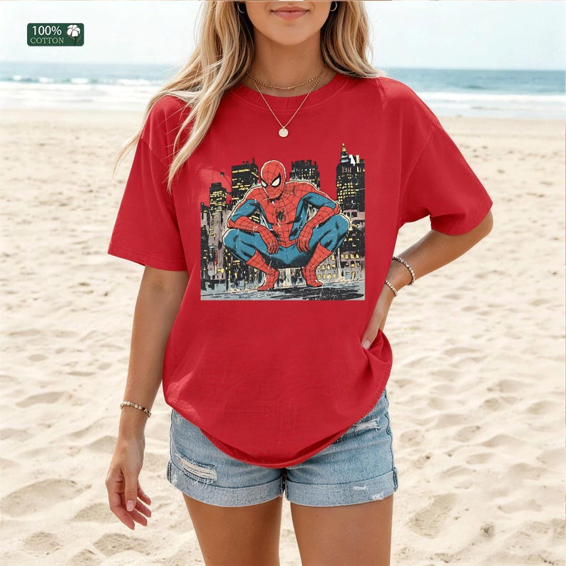 May include: A red short-sleeve t-shirt featuring a graphic print of the character Spider-Man in a crouching position against a dark city skyline background. The shirt has a crew neckline and a small square label in the corner stating 100% cotton.