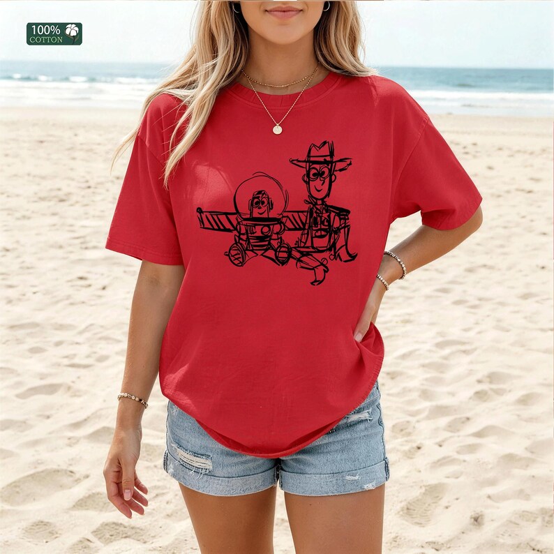 May include: A bright red short-sleeved t-shirt featuring a black line-art graphic of two cartoon characters, one wearing a space helmet and the other a cowboy hat. The shirt has a crew neckline and a small square label in the corner reading 100% COTTON.